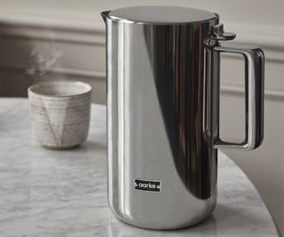 An Aarke kettle on a kitchen table beside a steaming cup of water.