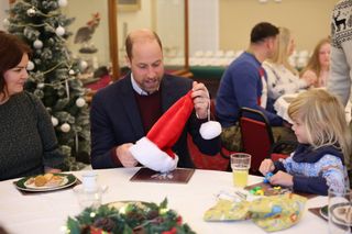 Prince William holding a santa hat sitting at a table with a little girl and mom with a tree behind them