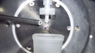 A droplet of metal squeezes out of a syringe in the center of the image; a tube that dispels water vapor is pointed at the droplet