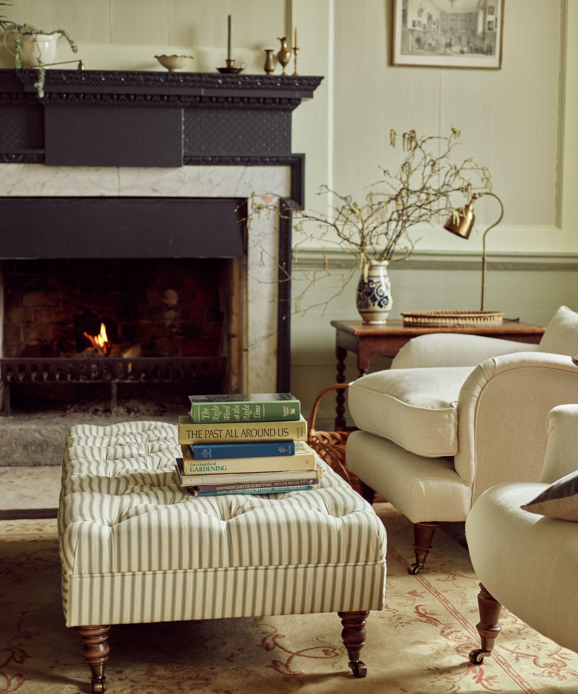 traditional living room painted in a soft neutral with wall panelling and a black fireplace with a striped ottoman