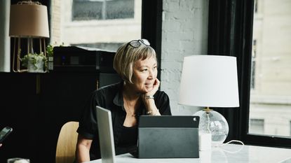 An older woman sits at her office desk, looking happily thoughtful.