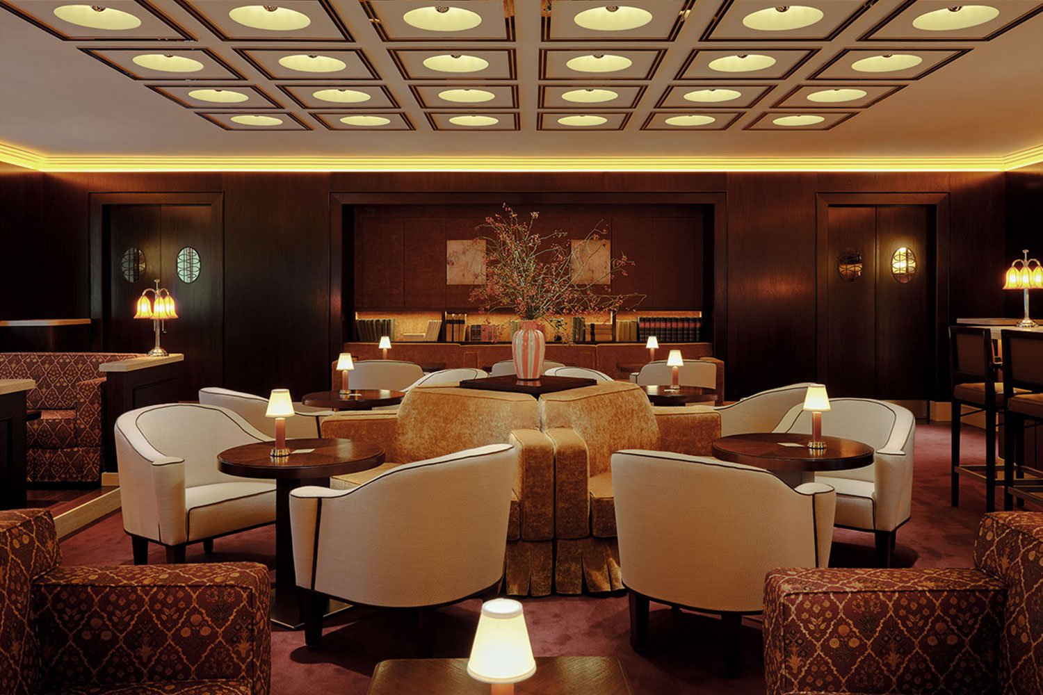 A cinematic cocktail lounge with coffered, back-lit ceilings, plush patterned textiles, and a concentric constellation of soft seating is brought to life by vintage table lamps.