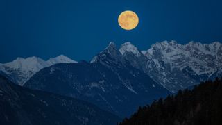 close up view of a perfectly full moon rising in the partly snow covered mountains. Majestic mountain peaks are visible. The sky is intensely blue with no clouds.