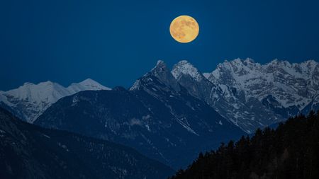 close up view of a perfectly full moon rising in the partly snow covered mountains. Majestic mountain peaks are visible. The sky is intensely blue with no clouds.