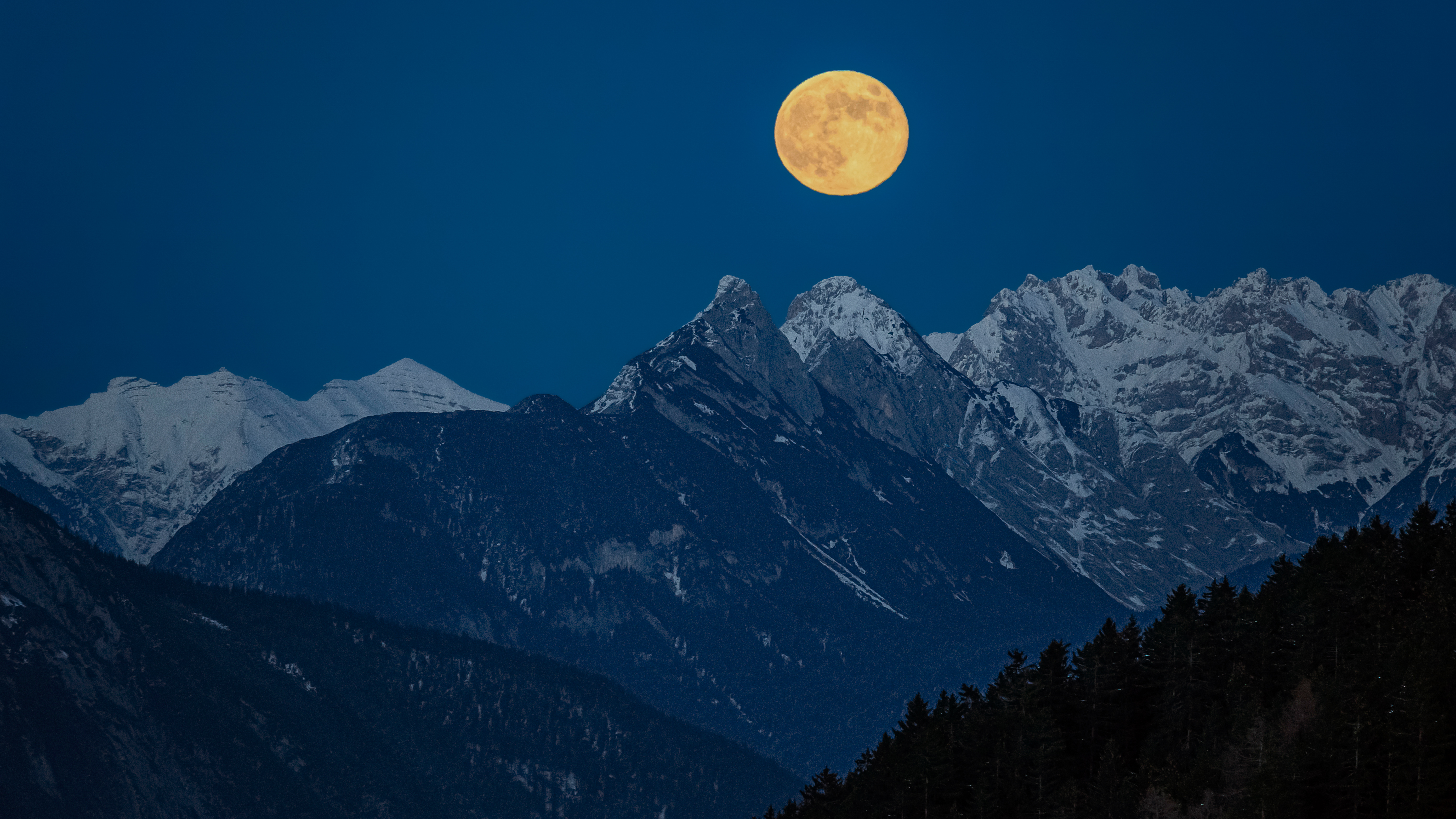 close up view of a perfectly full moon rising in the partly snow covered mountains. Majestic mountain peaks are visible. The sky is intensely blue with no clouds.