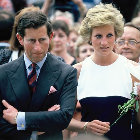 King Charles crosses his arms and wears a suit and striped tie while Princess Diana wears a dress with a white top and a black bodice while holding roses