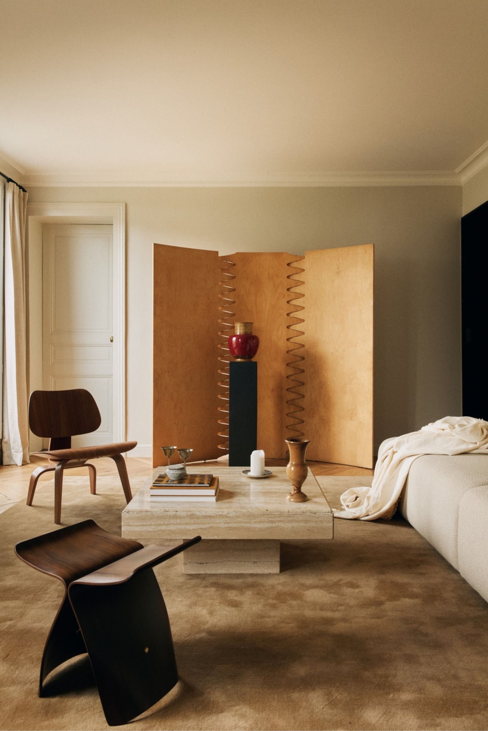 Living room with a wooden lounge chair, paper floor lamp, sculptural wooden screen, black pedestal, decorative vases and a beige rug on herringbone wood flooring.
