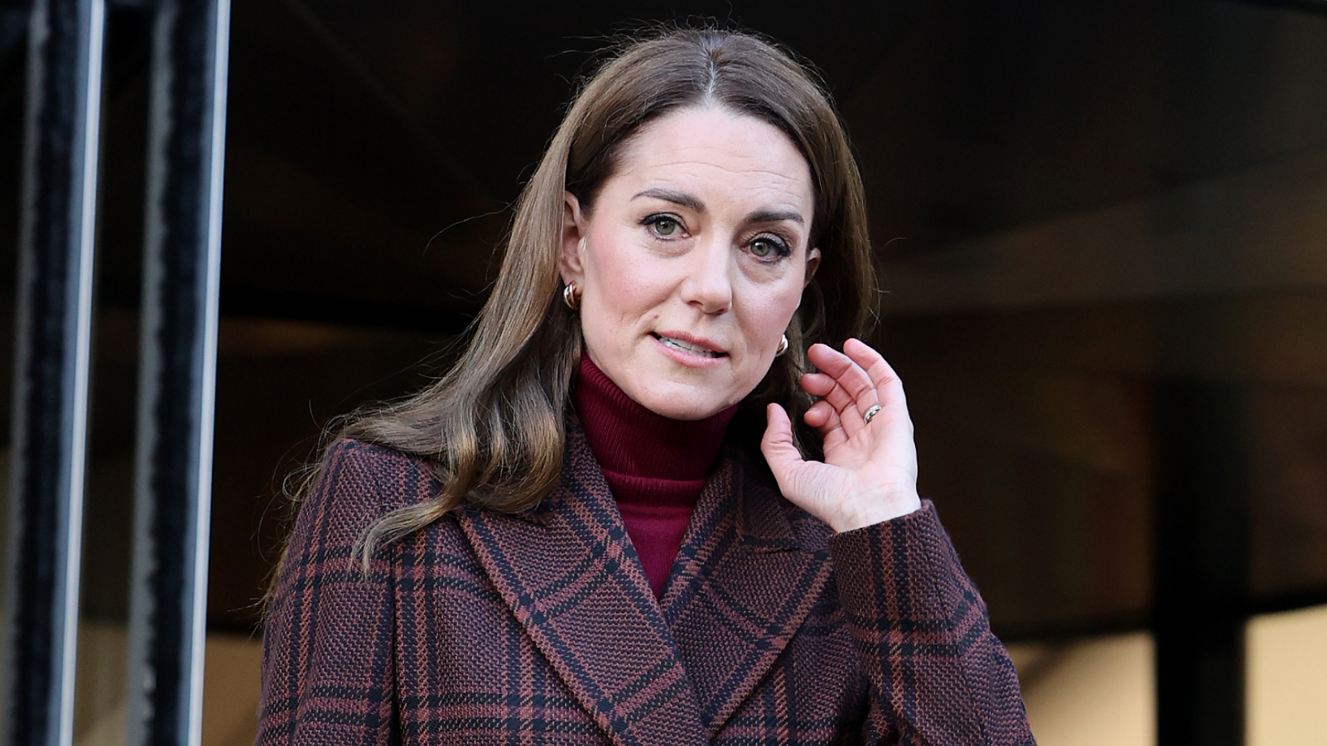 Kate Middleton has long brown wavy hair and wears a brown blazer with jeans, while wearing a tartan overcoat and heels