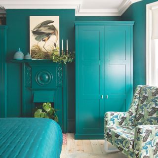 Teal bedroom with teal paint, teal bedding, and a teal wardrobe. There's a fabric chair in a leaf print sitting in front of the wardrobe