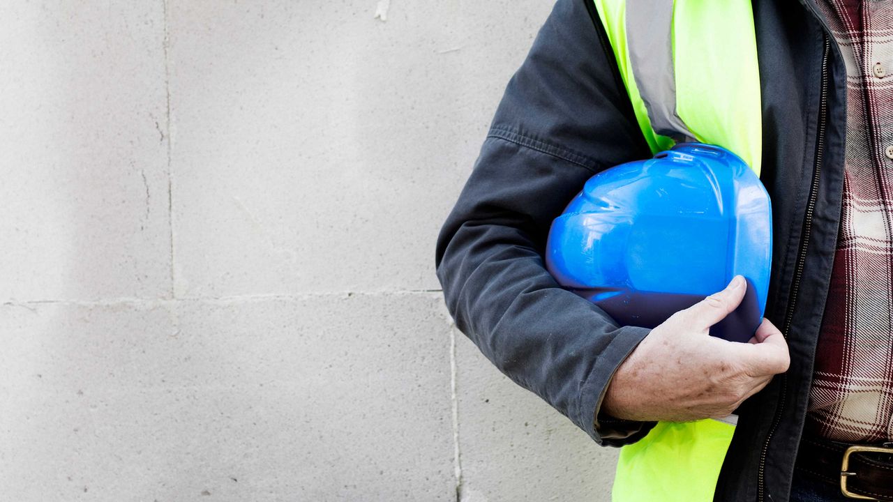 A man wearing a safety vest holds a construction hardhat under his arm.