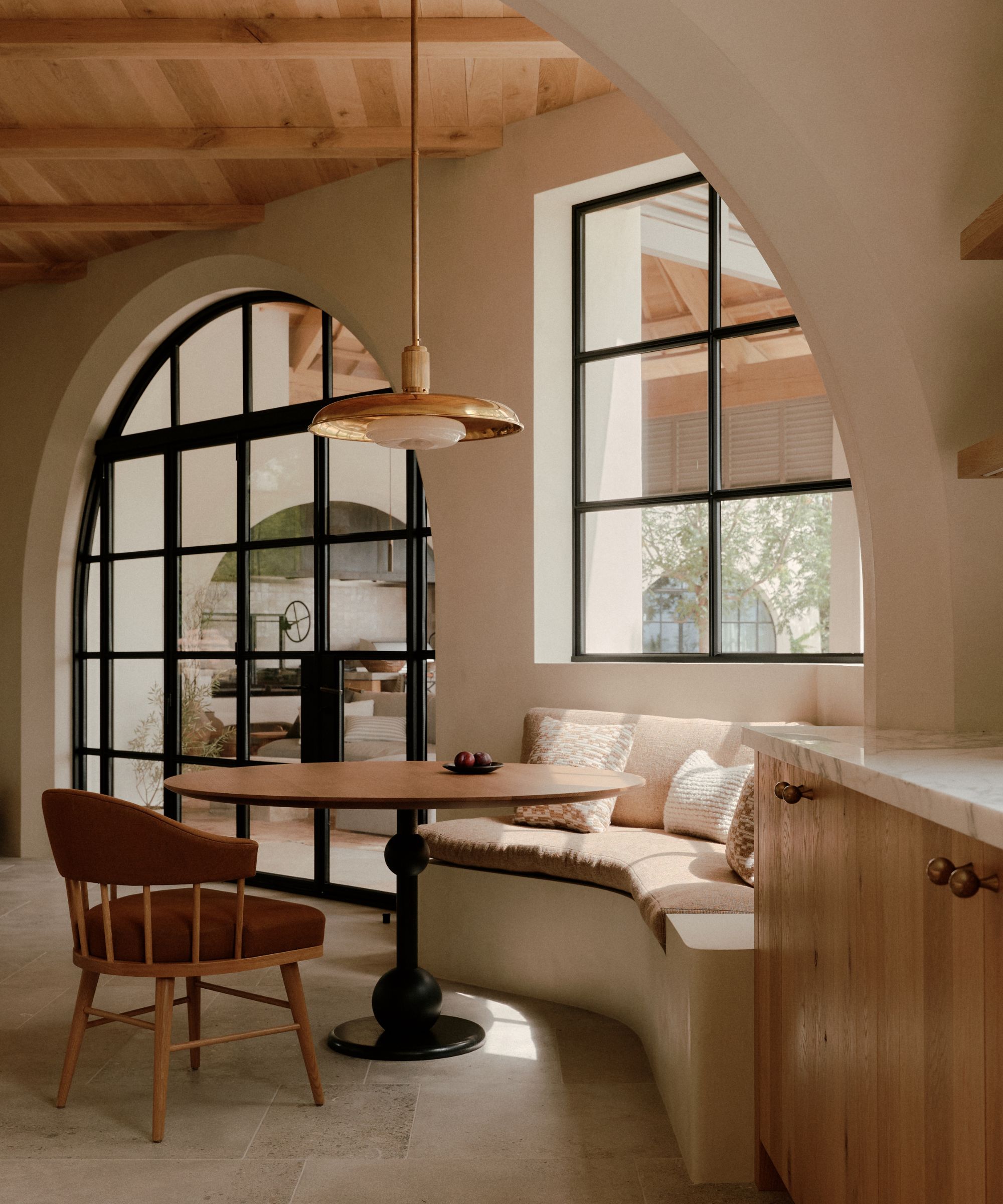 A sunlit breakfast nook featuring a curved, cream-colored built-in banquette and a round wooden pedestal table. A brass pendant light hangs above, framed by a large arched window and black steel-framed glass doors