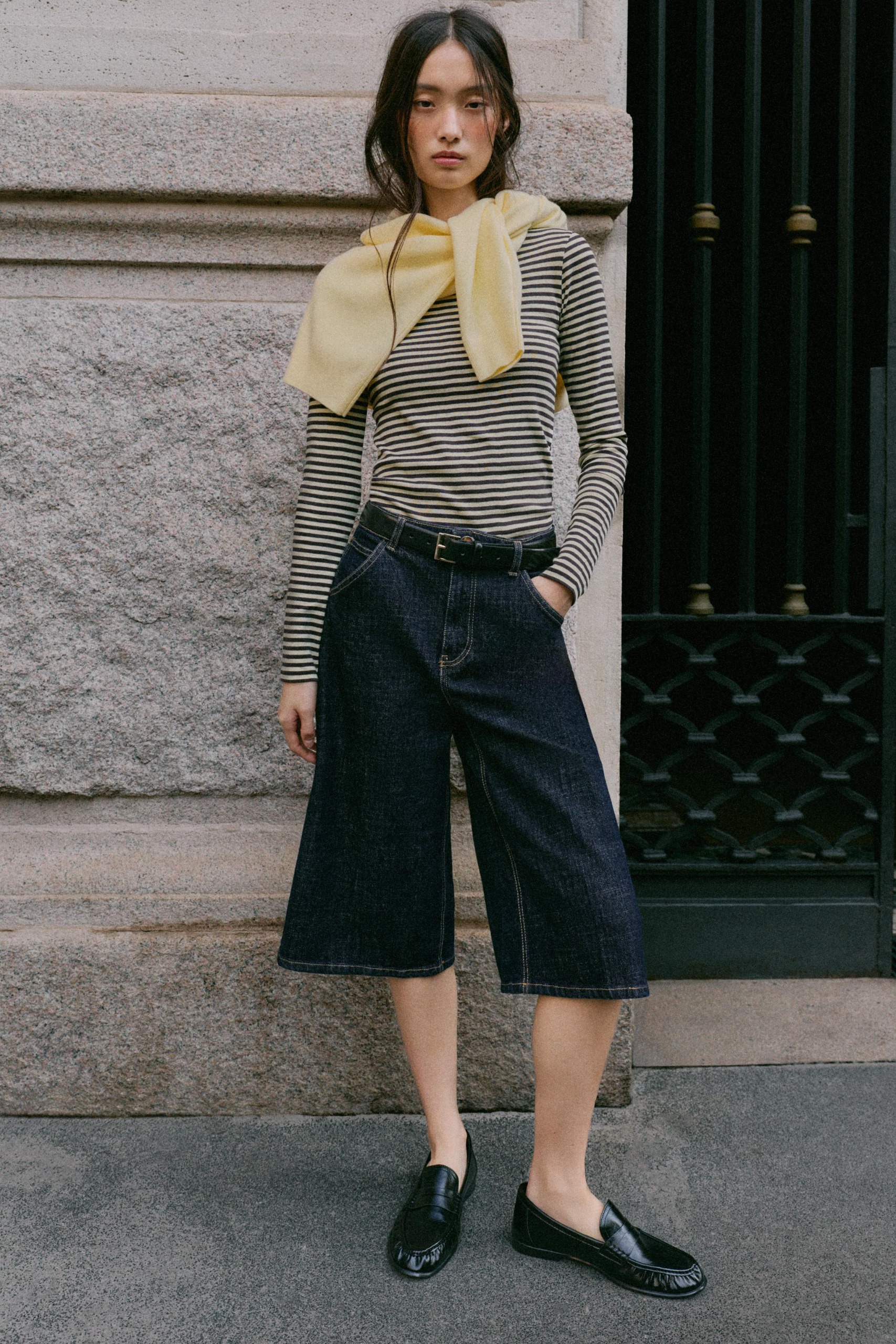Woman wears black and white striped shirt, long denim short/culottes, and black loafers.