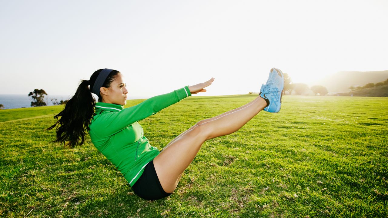 A woman in a sports jacket, sneakers and shorts does an abs workout in a grassy field. She is sitting on the floor, with her legs and torso elevated creating a &#039;V&#039; shape, and her hands reaching towards her toes. In the distance we can see trees.