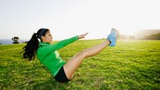 A woman in a sports jacket, sneakers and shorts does an abs workout in a grassy field. She is sitting on the floor, with her legs and torso elevated creating a 'V' shape, and her hands reaching towards her toes. In the distance we can see trees.