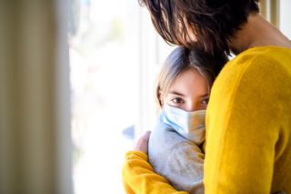 Vaccine misinformation: Small schoolgirl with face mask and unrecognizable mother indoors at home, hugging.