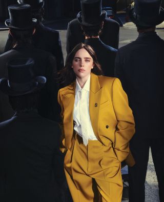 Billie Eilish wearing a gold suit and white shirt and tie standing behind men in tophats