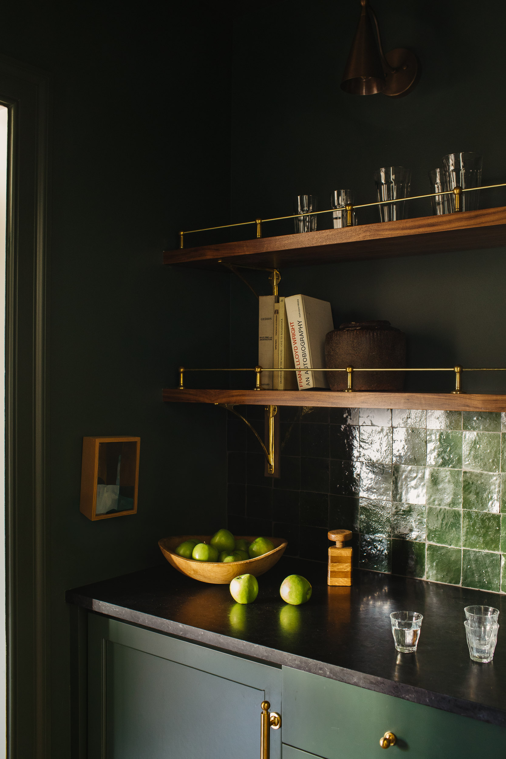 A dark green bar area with gallery shelves, black countertops, and a green tile backsplash. There is a wooden bowl full of limes and shot glasses on the counter.