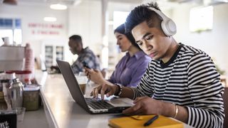 A man sits at the counter top of a coffee shop working on his laptop. In the background, other people are also working. He's wearing headphones to block out distracting sounds to aid concentration