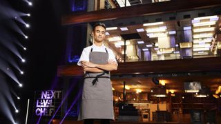 Contestant Ronan in white top and grey apron in front of the kitchen in Next level Chef