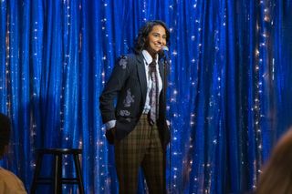 Amit Kaur as Bela, onstage speaking into a mic in front of a stool and sparkling blue curtain, in 'The Sex Lives of College Girls' season 4.