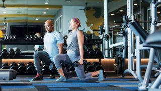 Woman and man perform lunges in gym holding dumbbells by their sides