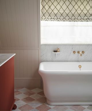 A panelled bathroom with a large white porcelain bath with gold faucets, a red and white chequered tiled floor and a red sideboard with marble countertop and marble backsplash behind the bath below a bright window with a cream and green geometric patterned blind