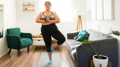 A woman stands practicing yoga in a living room. She is standing on her right leg, with her left leg bent and her left foot placed on the side of the standing lower leg. Her hands are held together at her chest as she looks forward. Behind her we see a couch, chair, low coffee table, decorative shelving and a large lamp.