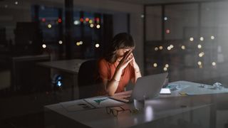 Woman late at night working from office desk with head in hands, representing burnout from work
