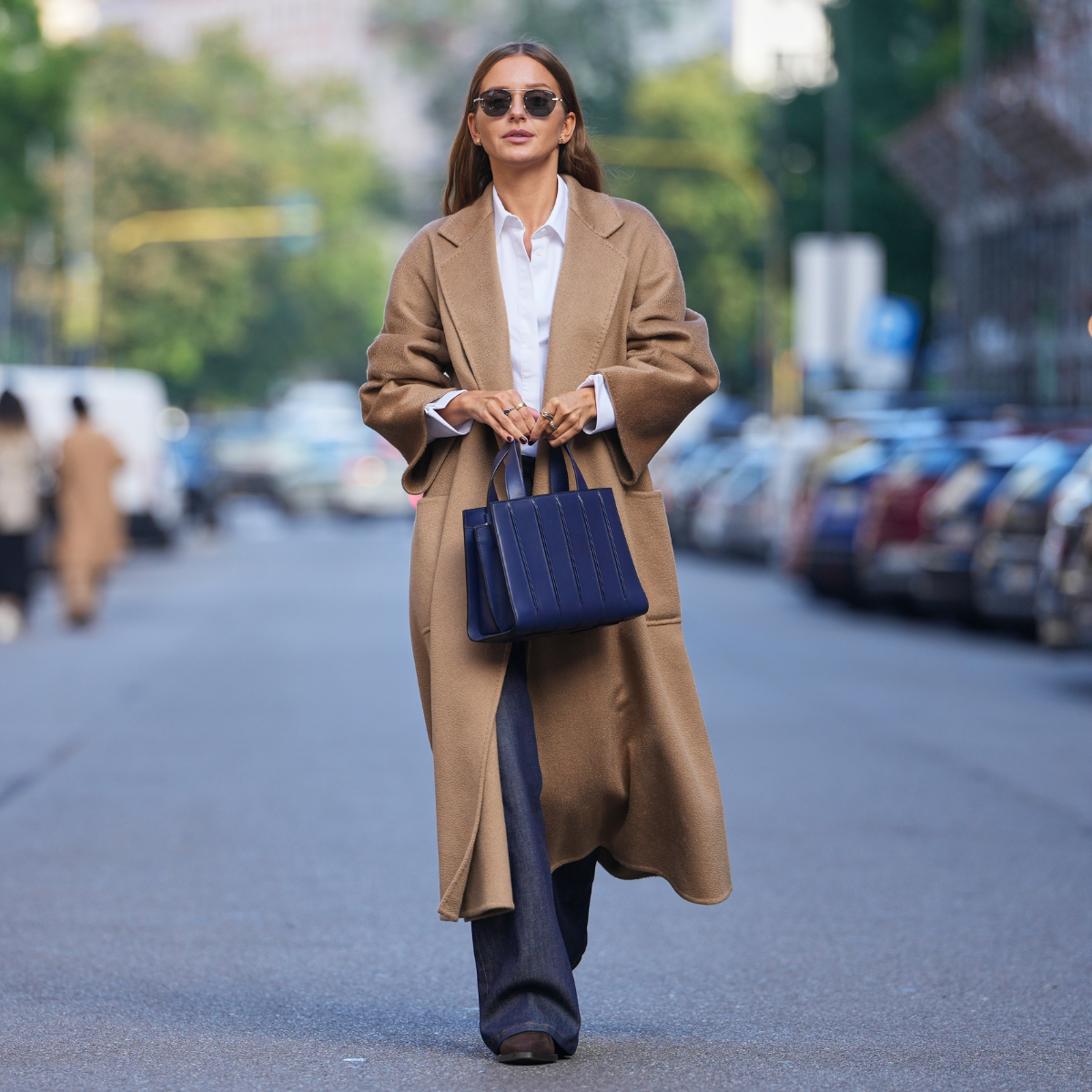 Diane Batoukina wears long straight brown hair with a center part, round black sunglasses, gold rings, a navy blue leather tote bag, a camel wool long coat, a white cotton button-up shirt, dark indigo denim wide-leg jeans, black leather boots shoes with a low block heel, outside Max Mara, during Milan Fashion Week 