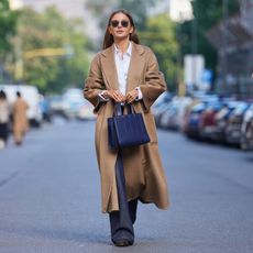 Diane Batoukina wears long straight brown hair with a center part, round black sunglasses, gold rings, a navy blue leather tote bag, a camel wool long coat, a white cotton button-up shirt, dark indigo denim wide-leg jeans, black leather boots shoes with a low block heel, outside Max Mara, during Milan Fashion Week