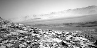 A photo of Mars taken by the Curiosity rover shows a bleak landscape of hills and hazy crater mountains.