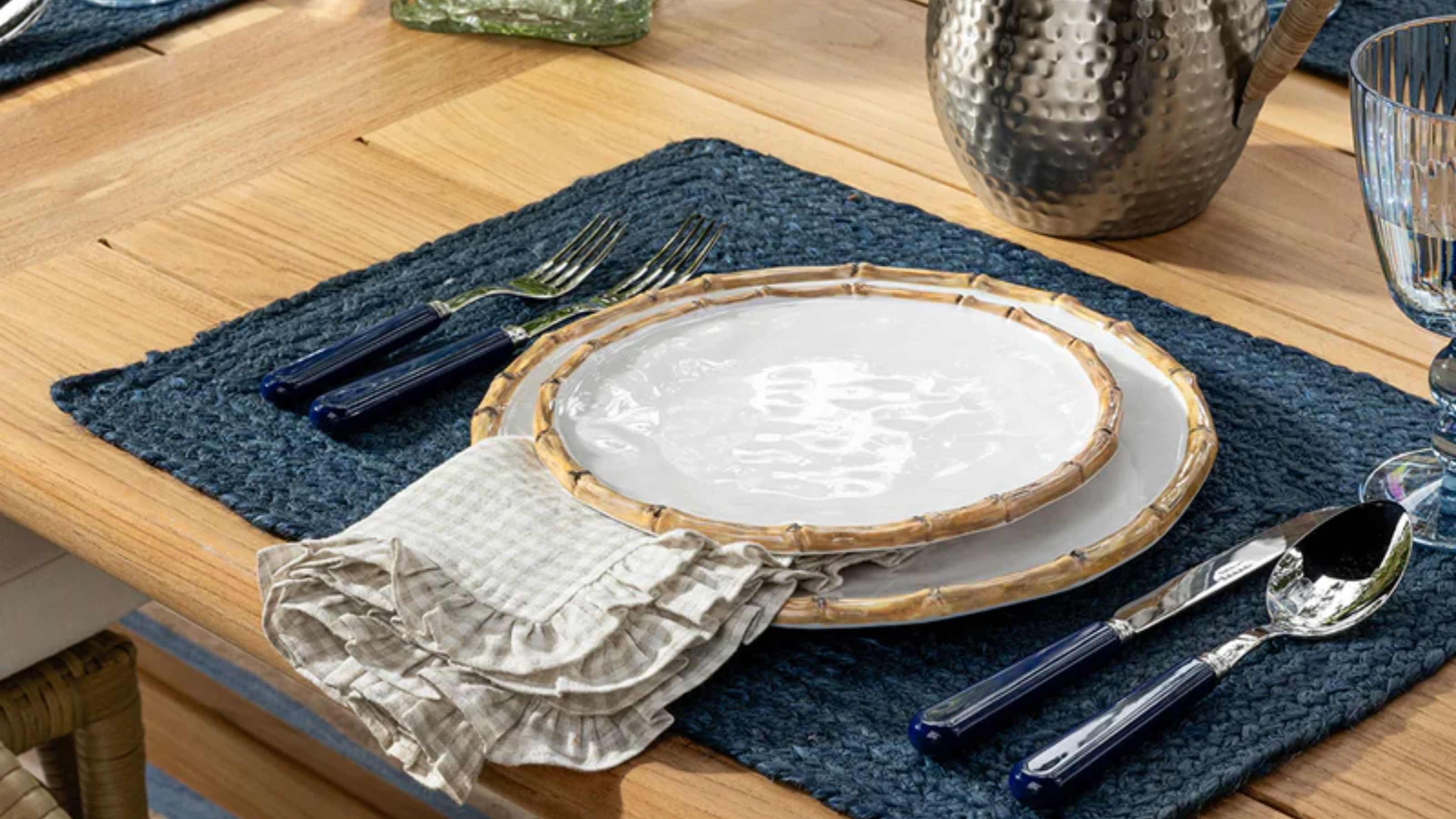 Bamboo plates styled alongside gingham napkin linen, navy placemat, and coordinating navy flatware atop of a wooden dining table