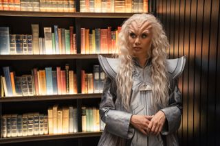 a humanoid alien with long blonde hair stands next to a bookshelf