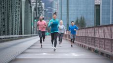 Four runners run across bridge and towards camera, there are tall office buildings in the background