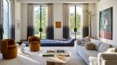 A bright living room with two-toned ombre curtains by tall windowed doors by a blue geometric floor sofa beside ochre sofa armchairs and a long cream couch by a potted tree, with a wood coffee table in the middle of the room