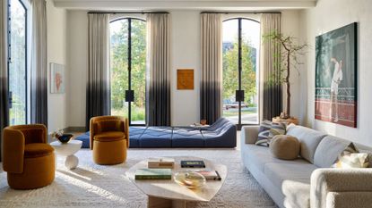 A bright living room with two-toned ombre curtains by tall windowed doors by a blue geometric floor sofa beside ochre sofa armchairs and a long cream couch by a potted tree, with a wood coffee table in the middle of the room