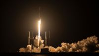 The bright orange-white plume from a rocket lights up its launch pad as it lifts off into the darkness of an early morning sky.