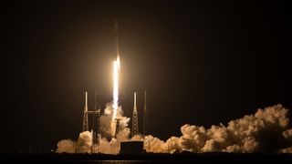 The bright orange-white plume from a rocket lights up its launch pad as it lifts off into the darkness of an early morning sky.