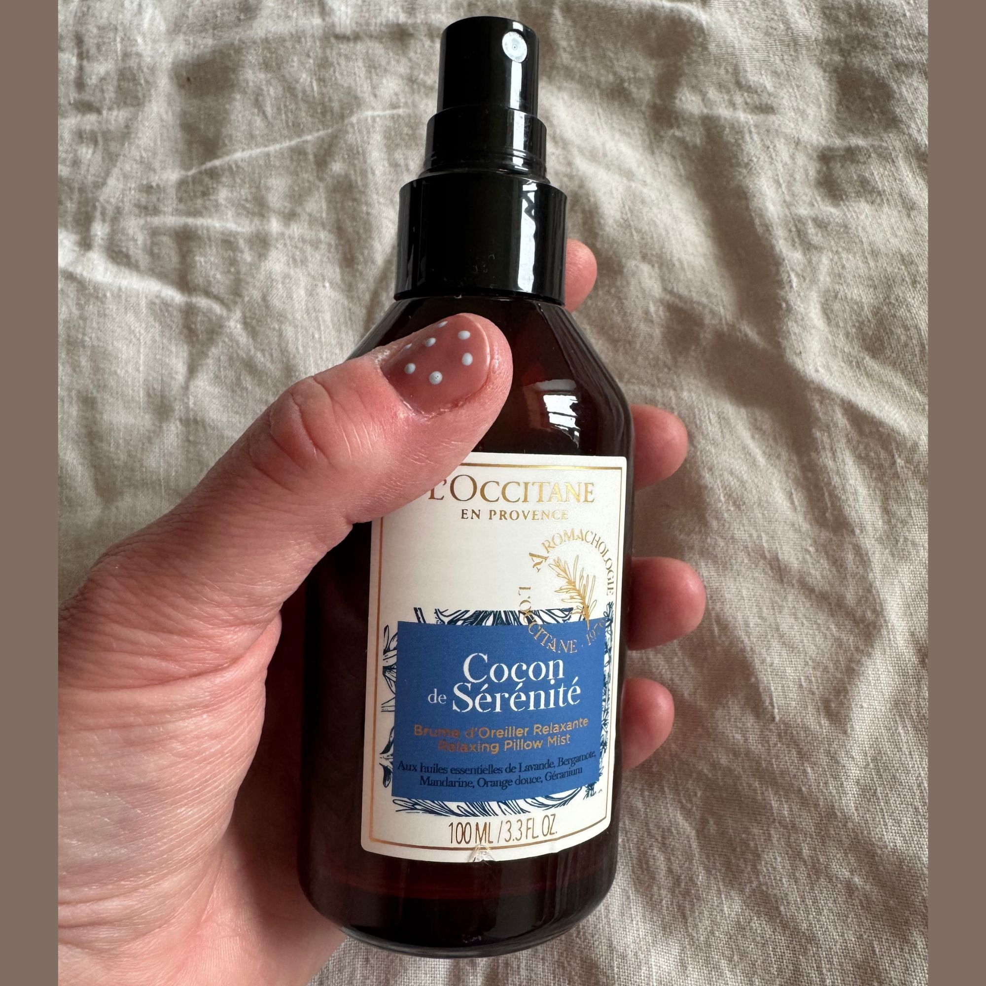 A bottle of L'occitane Cocon De S&amp;eacute;r&amp;eacute;nit&amp;eacute; Pillow Mist pictured in the writer's hand with her bed visible in the background