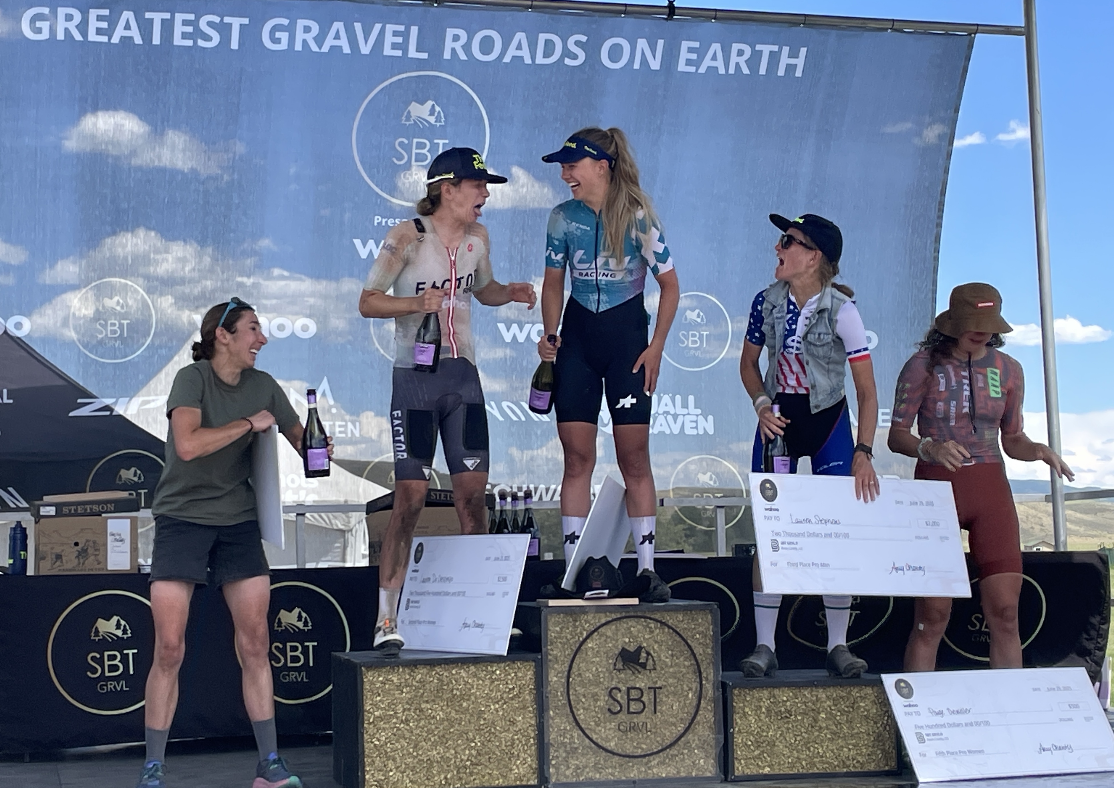 Melisa Rollins (centre) celebrates on the SBT GRVL podium as the women's elite winner, sharing a shower of sparkling wine alongside Lauren De Crescenzo, Lauren Stephens, Sarah Lange and Paige Onweller