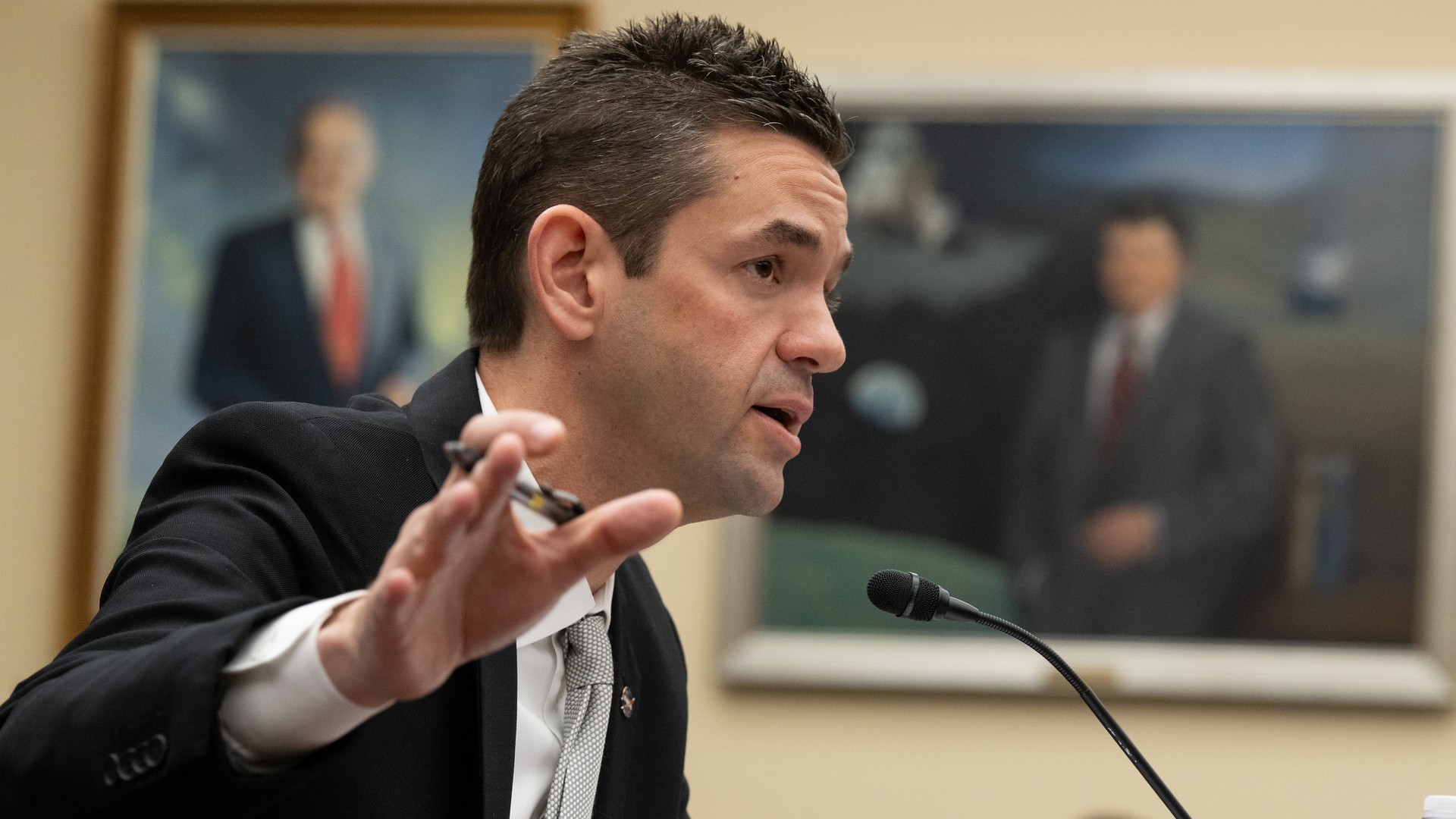 a man in a black suit speaks into a microphone in a large hearing room