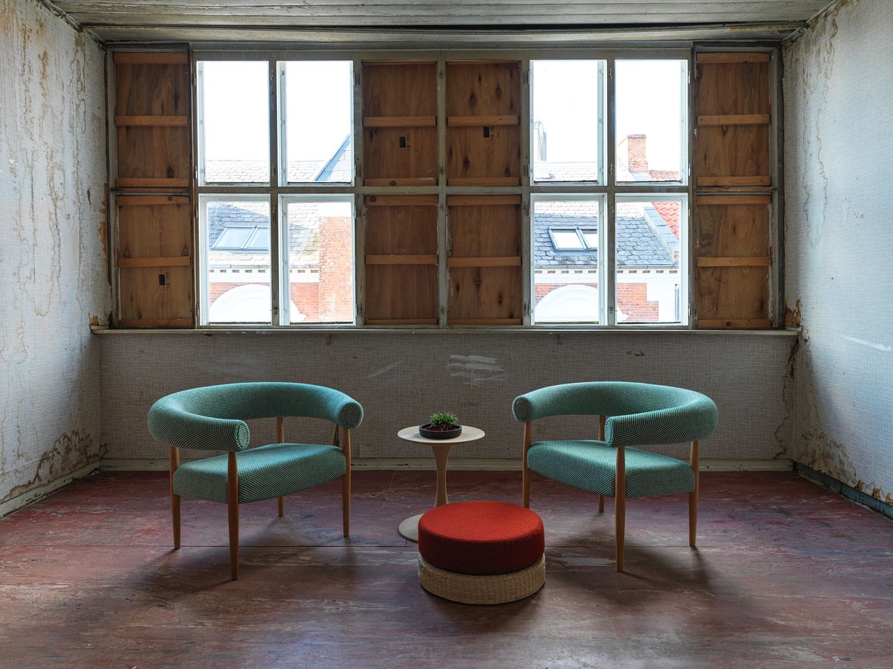 A sitting area with two wooden blue cushioned chairs, a red foot stool and and round coffee table with a potted plant on it in front of two windows.