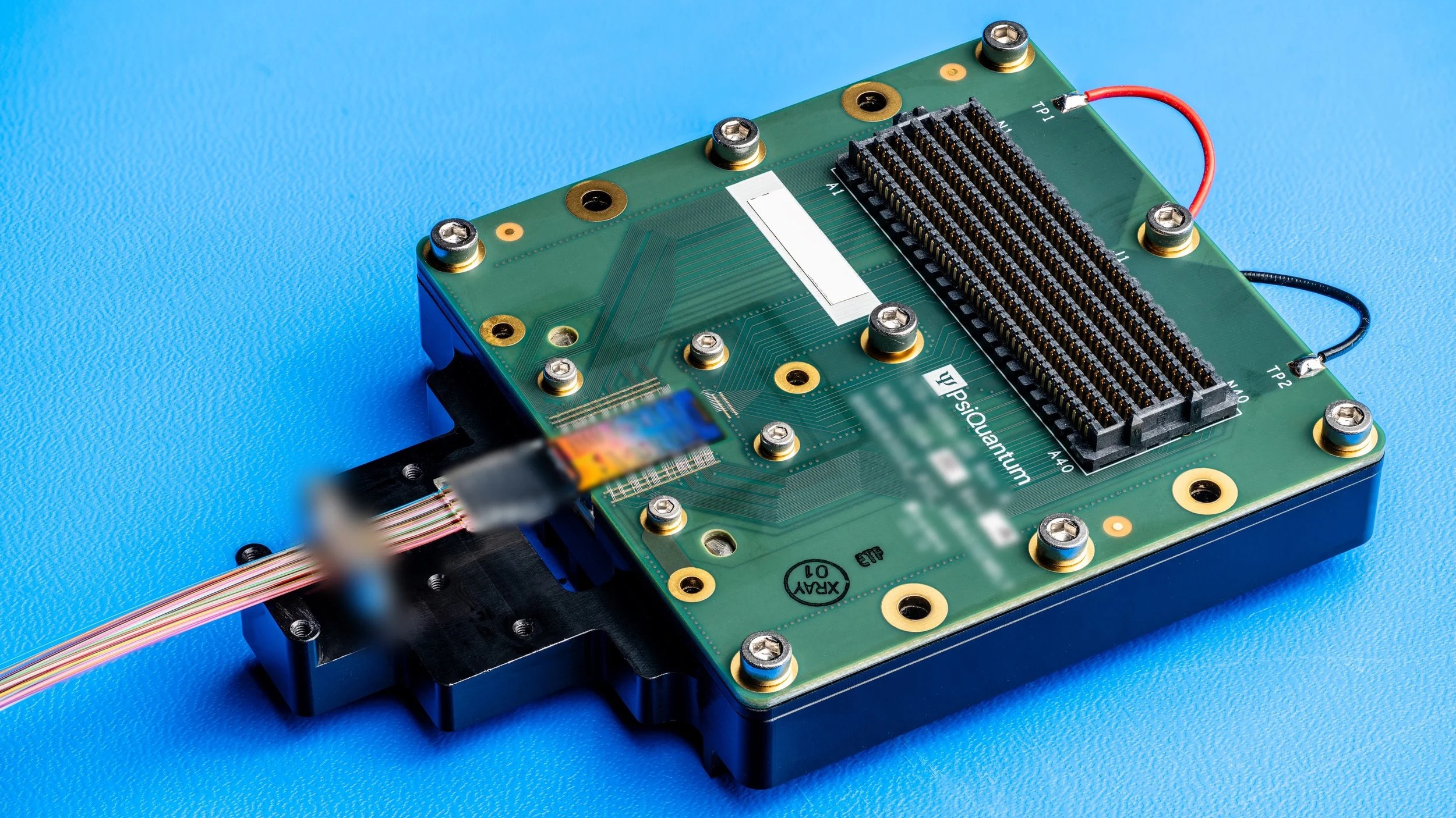 A green PsiQuantum circuit board module with a photonic chip and a ribbon of optical fibers attached, on a blue background.