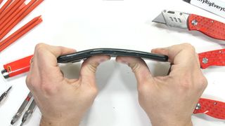 A screenshot from JerryRigEverything's durability test of the OnePlus 10 Pro, showing the phone's damage from a bend test. The phone is seen side-on from above, with the body bent at a slight angle away from the host's hands
