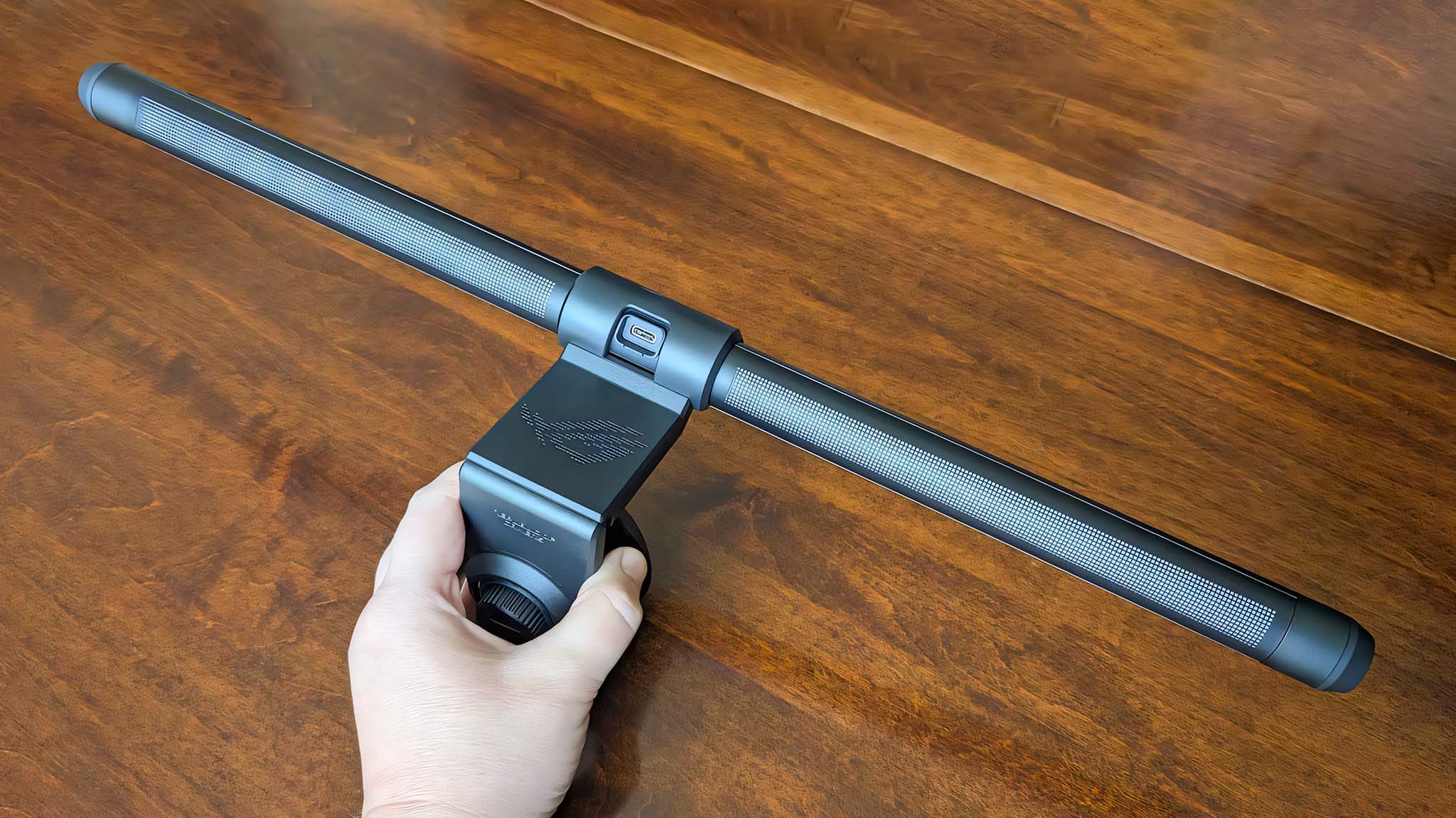 A hand holding the ASUS ROG Aura Monitor Light Bar above brown wooden table.