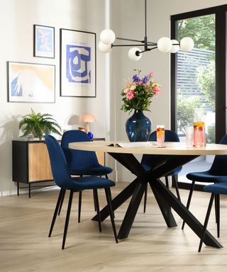 A dining room with white walls with three wall art prints, a hanging black chandelier, a round wooden dining table with black legs and blue curved seats underneath it, and beige flooring underneath it