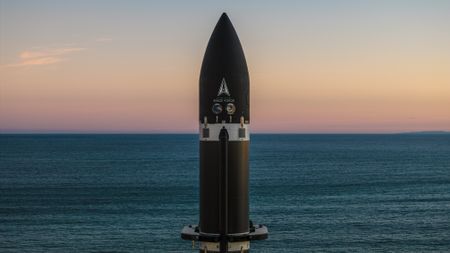 The top of a black rocket stands against a background of rich sea parted in the center by the horizon, and a cool, orange-hued sky. 