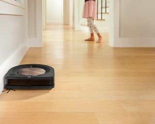 iRobot S9 next to skirting board on laminate wooden floor with female child wearing spotted leggings in background