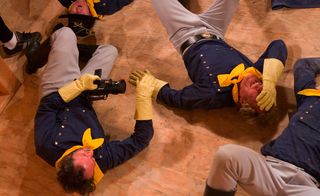 A video still of people in gray, deep blue, and yellow uniforms are lying on the floor. They seem like they are in the middle of a scene. One person is holding a camera and is filming the rest.