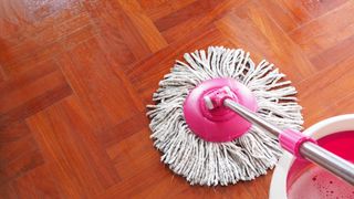 A round mop head on a wooden floor next to a bucket of soapy water